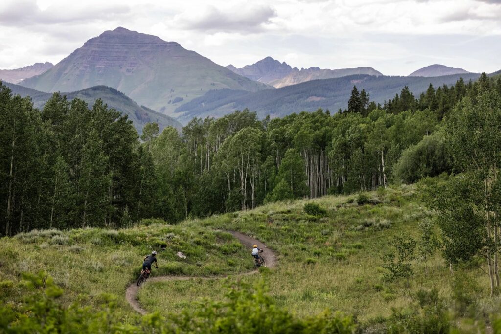 2 mountain bike riders descend a flow train with big mountains in the distance